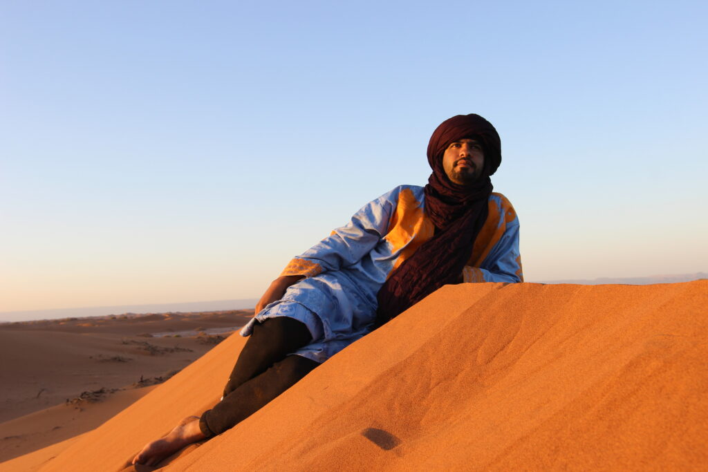 In the picture, a nomad stands gracefully atop a vast expanse of golden sand dunes, their silhouette striking against the endless sky. The nomad is adorned in traditional attire, featuring a long, flowing robe that gently flutters with the desert breeze. Their head is wrapped in a distinctive turban, its layers and folds providing protection from the relentless sun and blowing sand. The colors of the turban and robe are earthy and muted, blending harmoniously with the desert landscape. The nomad's posture is one of quiet strength and resilience, embodying the spirit of a life lived in harmony with the harsh but beautiful environment. In the background, the rolling dunes stretch out to the horizon, creating a serene and timeless scene.